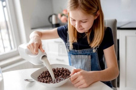 Photo for Sweet Girl Pours Milk Into Bowl With Dry Breakfast In Kitchen - Royalty Free Image