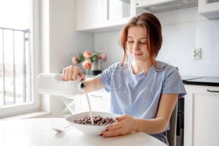 Téléchargez les photos : Adolescente sucrée verse du lait dans un bol avec petit déjeuner sec dans la cuisine - en image libre de droit
