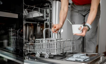 Photo for Young Woman Is Loading Spoons Into Dishwasher In Close-Up View - Royalty Free Image