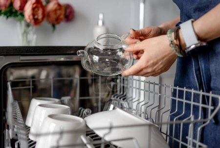 Weibliche Hände halten eine transparente Teekanne gegenüber einer Spülmaschine, die sauberes Geschirr anrichtet