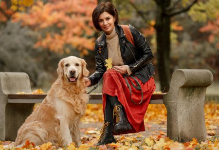 Téléchargez les photos : Une jeune femme assise sur un banc dans un parc d'automne et caressant un chien Golden Retriever - en image libre de droit