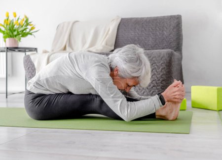 Téléchargez les photos : Femme athlétique âgée se plie tout en étant assis sur un tapis de yoga à la maison sur le sol dans une pièce lumineuse avec des fleurs - en image libre de droit