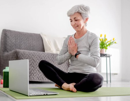 Téléchargez les photos : Agréable femme de 65 ans salue son entraîneur par ordinateur portable pour une séance de sport en ligne à la maison sur un tapis de yoga - en image libre de droit
