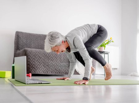 Foto de Agradable mujer de 70 años realiza ejercicios complejos con entrenador a través de computadora portátil, haciendo yoga en línea en casa - Imagen libre de derechos