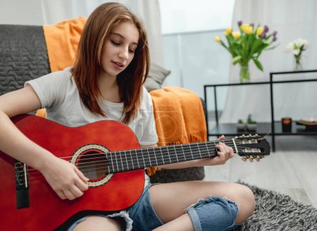 Téléchargez les photos : Adolescente pratiquant la guitare à la maison assise sur le sol. Jolie guitariste avec instrument de musique - en image libre de droit