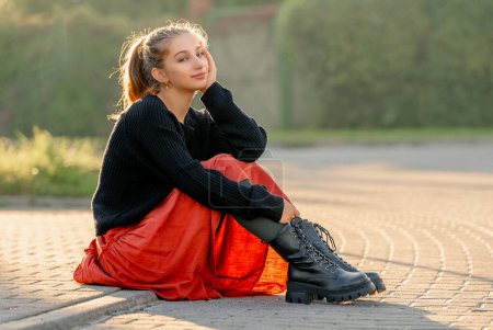 Téléchargez les photos : Belle adolescente en jupe rouge assise à l'extérieur dans la rue. Jolie adolescent modèle posant dans des vêtements à la mode - en image libre de droit