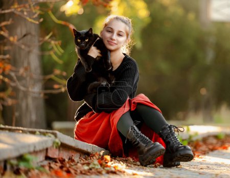 Téléchargez les photos : Jolie fille en jupe rouge étreignant chat noir à l'extérieur dans la rue avec des feuilles d'automne. Belle adolescente modèle assise avec un animal félin au parc - en image libre de droit