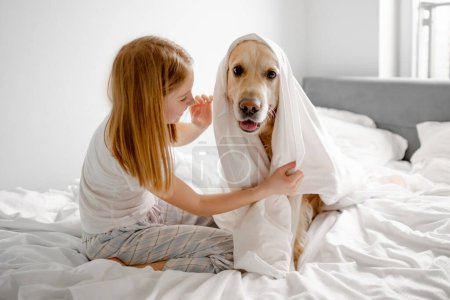 Foto de Chica juega con Golden Retriever en la cama, cubriéndolo con manta en la habitación brillante por la mañana - Imagen libre de derechos