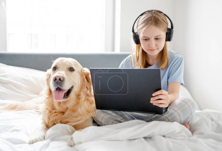 Téléchargez les photos : Fille est assise dans le lit avec ordinateur portable, écouteurs et Golden Retriever chien dans la chambre lumineuse le matin - en image libre de droit