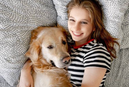 Téléchargez les photos : Jolie fille étreignant chien golden retriever et souriant couché au lit. Adolescent heureux avec chien de race labrador animal de compagnie reposant ensemble - en image libre de droit