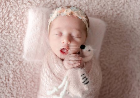 Téléchargez les photos : Fille nouveau-né enveloppé dans une couverture avec un jouet de flamant rose dans ses mains bâille lors d'un Photoshoot nouveau-né professionnel - en image libre de droit