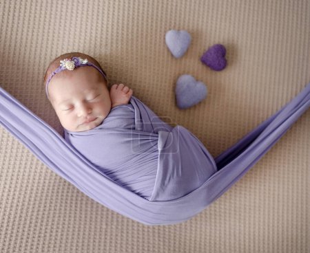 Téléchargez les photos : Fille nouveau-né enveloppé dans l'hamac dort dans des tons lilas pendant la session professionnelle de photo de nouveau-né - en image libre de droit