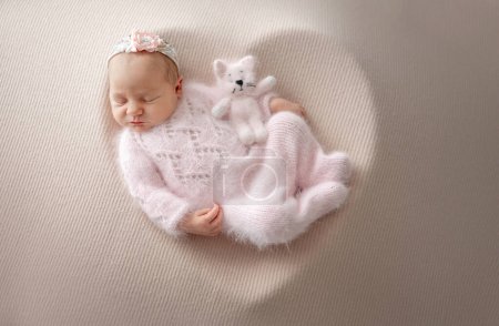 Neugeborenes Mädchen im rosafarbenen Anzug mit Spielzeugkatze schläft während des professionellen Fotoshootings für Neugeborene in einer herzförmigen Schüssel aus Holz