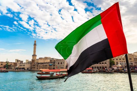 Photo for Focus on a flag of  United Arab Emirates flag against Abra - old traditional wooden boat and Grand Bur Dubai Masjid Mosque on the bay Creek in Dubai - Royalty Free Image