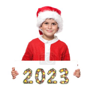 Foto de Boy Santa Claus sostiene un cartel de navidad y un número 2023 hecho por ramas de árbol de navidad aisladas sobre fondo blanco - Imagen libre de derechos
