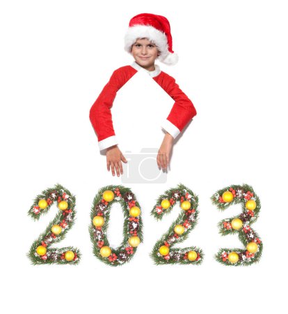 Téléchargez les photos : Garçon Père Noël tient une affiche de Noël et 2023 nombre fait par branches d'arbre de Noël isolé sur fond blanc - en image libre de droit