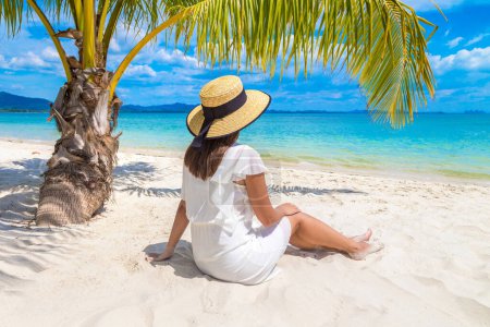 Foto de Joven hermosa mujer con vestido blanco y sombrero de paja está sentado debajo de la hoja de palma en una playa tropical - Imagen libre de derechos