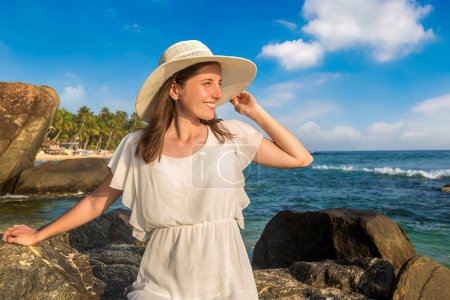 Téléchargez les photos : Portriat de jeune belle femme assise sur une pierre portant un chapeau et une robe blanche séjournant sur une plage tropicale près de la mer - en image libre de droit
