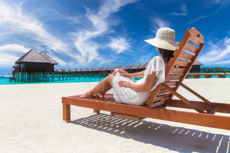 Téléchargez les photos : Belle femme se détendre dans un transat à la plage tropicale de luxe dans une journée d'été ensoleillée - en image libre de droit