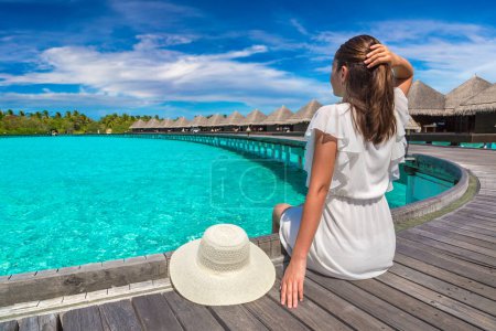 Photo for Beautiful young woman in front of water luxury villas sitting on the tropical beach jetty (wooden pier) in Maldives island - Royalty Free Image