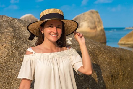 Téléchargez les photos : Femme voyageur en robe blanche et chapeau de paille à la belle plage tropicale rocheuse dans une journée d'été - en image libre de droit