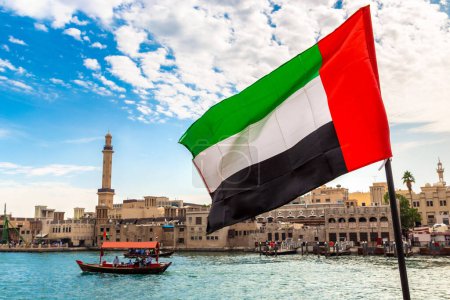 Foto de Enfócate en una bandera de los Emiratos Árabes Unidos contra Abra: viejo barco de madera tradicional y la mezquita Grand Bur Dubai Masjid en el arroyo Bay Creek en Dubai - Imagen libre de derechos
