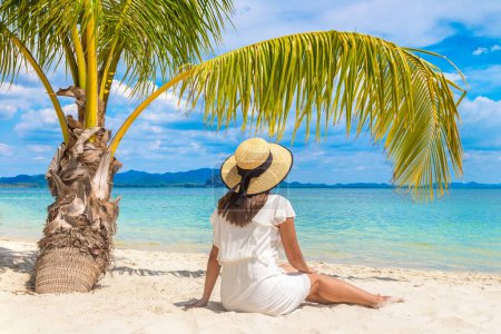 Téléchargez les photos : Jeune belle femme portant une robe blanche et chapeau de paille est assis sous la feuille de palme sur une plage tropicale - en image libre de droit