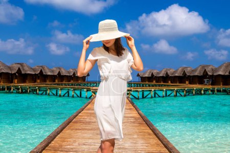 Foto de Hermosa mujer joven frente a villas de lujo de agua de pie en el embarcadero de playa tropical (muelle de madera) en la isla de Maldivas - Imagen libre de derechos