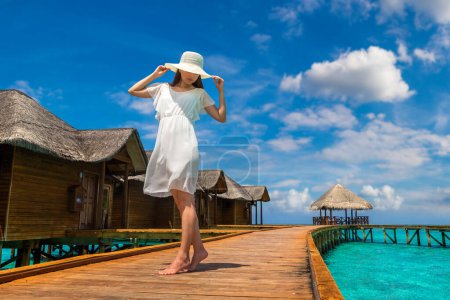 Foto de Hermosa mujer joven frente a villas de lujo de agua de pie en el embarcadero de playa tropical (muelle de madera) en la isla de Maldivas - Imagen libre de derechos