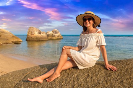 Foto de Mujer viajera con vestido blanco y sombrero de paja en la hermosa playa tropical rocosa en un día de verano - Imagen libre de derechos
