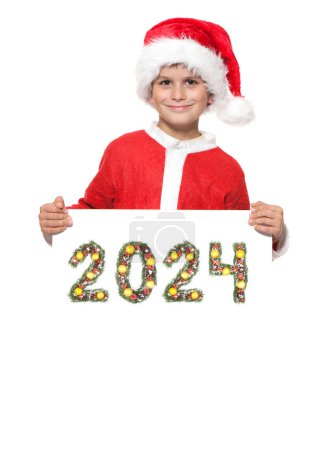 Téléchargez les photos : Garçon Père Noël tient une affiche de Noël et 2024 nombre fait par branches d'arbre de Noël isolé sur fond blanc - en image libre de droit
