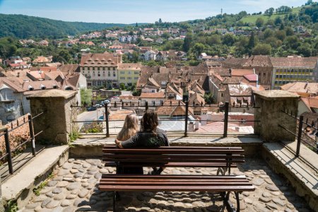 Photo for SIGHISOARA, ROMANIA - august 10, 2019:  Young couple sitting on a bench observes old streets  from a view point in  Sighisoara, Romania,  in well-preserved old town, listed by UNESCO as a World Heritage Site - Royalty Free Image