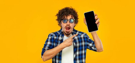 Foto de Design for Mobile App Advertisement. Man Showing Smartphone Screen On Yellow Studio Background. Smiling Guy on Ads Template. Cellphone Display Mockup - Imagen libre de derechos