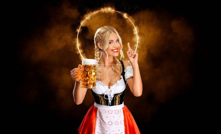 Foto de Retrato de media longitud de la joven rubia sexy con el pecho grande vistiendo dirndl negro con blusa blanca sosteniendo la taza de cerveza aislada sobre fondo oscuro - Imagen libre de derechos