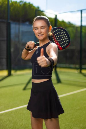 Téléchargez les photos : Joueur de tennis Padel avec raquette. Heureuse athlète fille souriante avec les pouces vers le haut sur le terrain à l'extérieur. Concept sportif. Téléchargez une photo de haute qualité pour la conception d'une application sportive ou d'un site Web - en image libre de droit