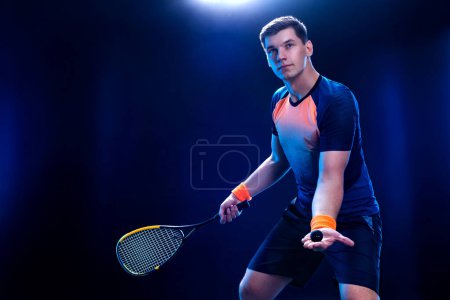 Foto de Jugador de squash en una cancha de squash con raqueta. Hombre atleta con raqueta en la cancha con colores neón. Concepto deportivo. Descargar una foto de alta calidad para el diseño de una aplicación deportiva o sitio de apuestas - Imagen libre de derechos