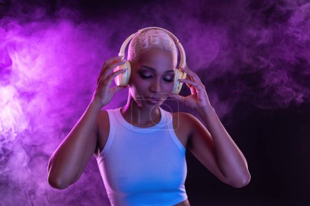 Photo for Hipster fashion girl, african american model wear headphones enjoy listen new cool music mix. Woman stand at purple studio background in trendy club purple party light with fog - Royalty Free Image