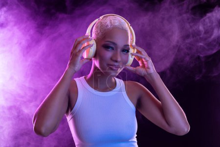 Téléchargez les photos : Hipster fille de la mode, modèle afro-américain porter des écouteurs profiter écouter nouveau mélange de musique cool. Stand de femme au fond de studio violet dans la lumière de fête violette club tendance avec brouillard - en image libre de droit