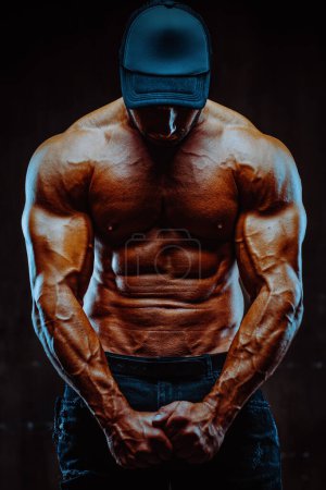 Téléchargez les photos : Young strong man bodybuilder in cap on dark background, dramatic colors - en image libre de droit