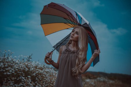 Téléchargez les photos : Young blond woman standing with umbrella in field with chamomile flowers, dark cold colors - en image libre de droit