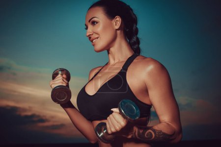 Photo for Young sports woman training with dumbbells on twilight sky background, tattoo on hand - Royalty Free Image