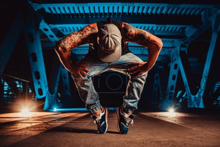 Téléchargez les photos : Jeune homme cool break danseur posant sur le pont urbain la nuit, tatouage sur le corps - en image libre de droit