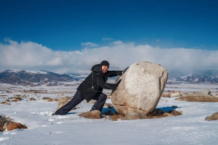Téléchargez les photos : Jeune homme émotionnel pousse un gros rocher sur fond de montagnes d'hiver - en image libre de droit