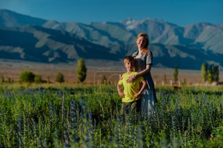 Foto de Mujer joven feliz con su hijo posando en un prado floreciente en el fondo de las montañas - Imagen libre de derechos