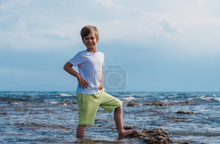 Téléchargez les photos : Portrait d'un garçon heureux de sept ans debout dans l'eau au bord de la mer en été - en image libre de droit