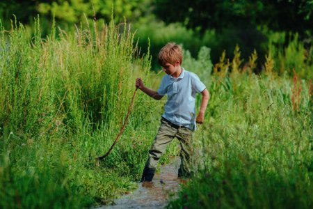 Foto de Niño en botas de goma con palo camina a lo largo de un arroyo poco profundo - Imagen libre de derechos