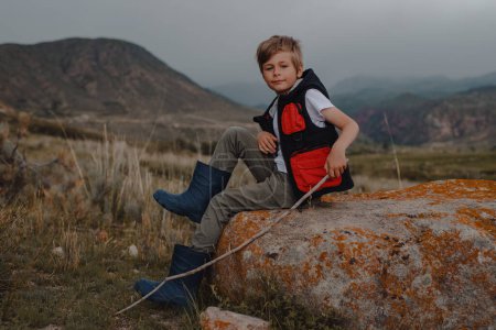 Foto de Niño excursionista con palo en un chaleco y botas de goma sentado en la piedra en las montañas - Imagen libre de derechos