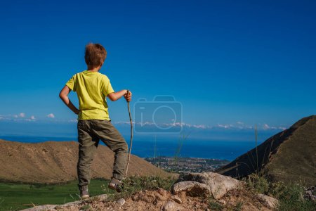 Photo for Boy hiker with stick looks at the lake shore from the top of the mountain - Royalty Free Image