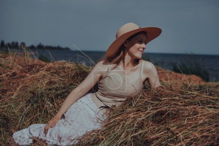 Téléchargez les photos : Belle jeune femme en chapeau reposant sur une botte de foin sur la rive du lac - en image libre de droit