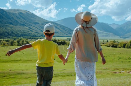 Foto de Feliz madre e hijo caminando a través de un prado verde en las montañas tomados de la mano - Imagen libre de derechos